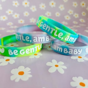 May include: Four colorful silicone bracelets with the text "Be Gentle, am Baby" on them. The bracelets are stacked on top of each other and have a tie-dye pattern.