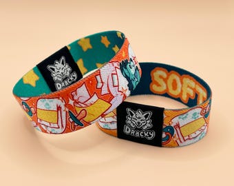 May include: Two elastic wristbands featuring an orange background with a repeating pattern of pacifiers and stylized shapes. Each band has a black fabric tag with a white graphic of a creature and the text Dracky. One band shows the word SOFT.