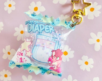 May include: A clear plastic keychain shaped like a bag of chips, with the words "Emergency Diaper" printed on it. The bag features a cartoon diaper design with stars and a pink cartoon character with a pacifier. The keychain has a gold star-shaped clasp.
