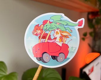 May include: A colorful sticker featuring a red car with three cartoon animal characters: a fox, a dragon, and a rabbit. The dragon is green with red accents, the fox is orange, and the rabbit is white and orange. The background is a light blue sky with clouds.