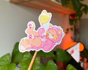 May include: A sticker featuring a cartoon fox in a pink dinosaur costume, holding a yellow cat-shaped balloon. The fox is lying down with its paws up, and has a pacifier. The sticker is on a wooden stick.