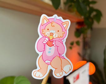 May include: A sticker featuring a cartoon character in a pink onesie with a fox-like hood, holding a red heart. The character has a pacifier and a bandage on its nose. The sticker has a white border and is held by a wooden stick.