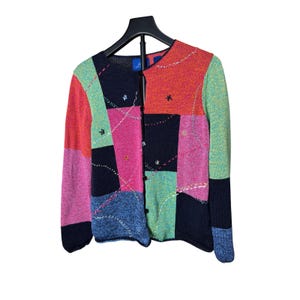 Op de afbeelding: Een kleurrijk patchwork-vest met een knoopsluiting. De trui heeft een mix van levendige kleuren, waaronder rood, roze, groen en blauw, gerangschikt in geometrische patronen. De mouwen zijn lang en het vest hangt aan een zwarte hanger.