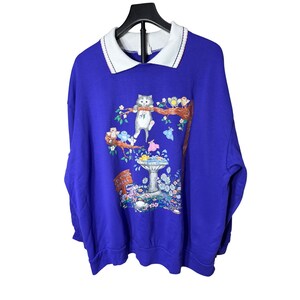 Pode incluir: Um moletom azul royal com gola branca com um gráfico de gato e pássaro. O gráfico inclui um gato pendurado em um galho de árvore, um bebedouro e o texto "Spring Carol".