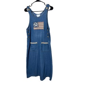 May include: A blue denim overall dress with an American flag patch on the front. The dress has two front pockets and decorative trim. The dress is hanging on a black hanger.