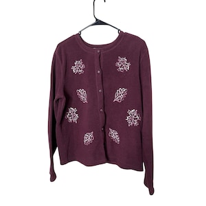 Peut inclure: Un cardigan bordeaux à manches longues et à boutons. Le pull présente des motifs de feuilles brodées blanches. Le vêtement est suspendu à un cintre noir.