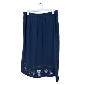 May include: Navy blue skirt with an elastic waistband, hanging on a white hanger. The hem features colorful embroidered Christmas-themed designs, including gifts, angels, and a Christmas tree.