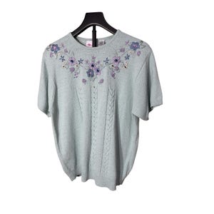 May include: A light blue short-sleeved sweater with floral embroidery in shades of purple and blue. The sweater features a cable knit design down the front. The garment is displayed on a black hanger against a white background.