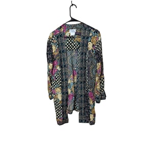 May include: A vintage open-front cardigan with a floral and paisley pattern in shades of green, purple, and gold. The long-sleeved garment is hanging on a black hanger against a white background. The cardigan has a small label.