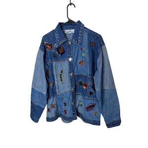 May include: A blue denim jacket with a patchwork design. The jacket is decorated with embroidered leaf and floral patterns in red, brown, and green. It has a button-up front and is suspended from a black hanger.