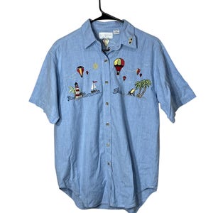 Op de afbeelding: Lichtblauwe shirt met korte mouwen en geborduurde strandscène. Voorzien van een vuurtoren, zeilboot, palmbomen en luchtballonnen in rood, geel en groen. Het shirt hangt aan een zwarte hanger.