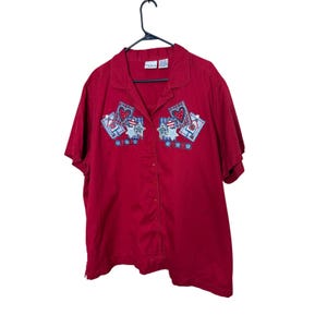May include: A red short-sleeved button-down shirt featuring a collar and embroidered patches on the chest. The patches display heart, star, and plaid square motifs in red, white, and blue colours, accented with small blue flower-shaped details.