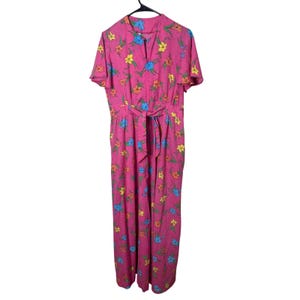 Puede incluir: Vestido rosa con estampado floral, mangas cortas y cinturón a juego. El vestido presenta un vibrante estampado de flores amarillas, azules y naranjas. Cuelga de una percha negra sobre un fondo blanco.