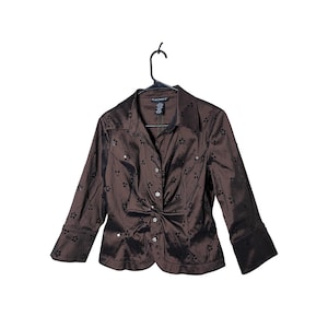 May include: A brown satin blouse with long sleeves and a button-down front. The blouse features a knotted design at the waist and is embellished with small, dark-colored floral accents. The blouse is hanging on a black hanger.