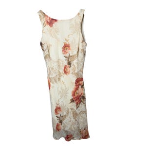 May include: A sleeveless, cream-colored dress with a floral pattern. The dress features large, red and pink roses with green leaves, set against a backdrop of beige paisley designs. The dress has a scoop neckline and a straight silhouette.