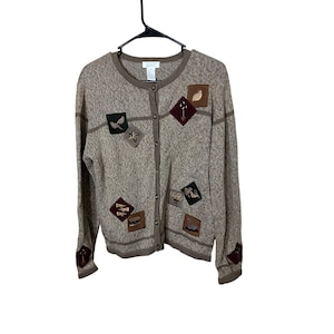 May include: A tan button-up cardigan sweater with various square patches featuring nature-themed designs. The patches include birds, leaves, and gardening tools. The sweater is hanging on a black hanger.