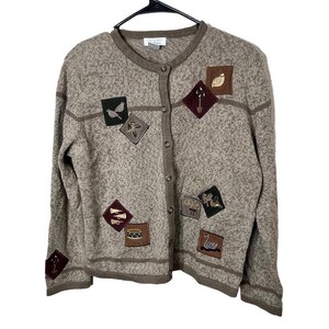 May include: A button-up cardigan in a speckled brown color, adorned with various square patches featuring embroidered designs. The patches include birds, musical instruments, and other nature-themed motifs. The sweater is hanging on a black hanger.
