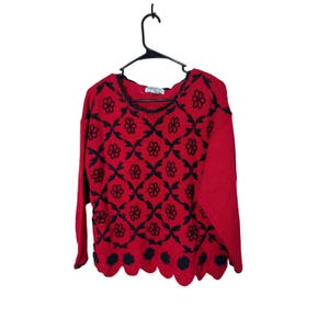 Op de afbeelding: Een rode trui met lange mouwen en een zwart bloemenpatroon over de voorkant. De trui heeft een geschulpte zoom met zwarte bloemaccenten. De trui hangt aan een zwarte hanger tegen een witte achtergrond.