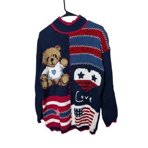 Puede incluir: Suéter azul marino con un gráfico de oso de peluche, corazón y diseño de bandera estadounidense. El suéter tiene un cuello alto y detalles en rojo y blanco. La palabra "Love" está escrita en la parte delantera.