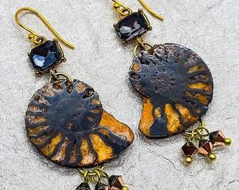 May include: A pair of gold earrings featuring a black and brown ammonite fossil with a dark blue gemstone accent. The earrings have a dangling design with small gold beads.
