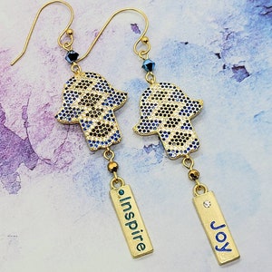 May include: Gold-toned earrings featuring Hamsa hand pendants with blue and black accents. Each earring has a rectangular charm with the word "inspire" or "joy" in blue. The earrings have a dangling design with small beads and a crystal accent.