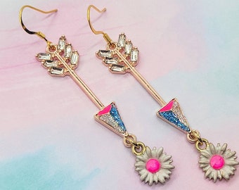 May include: A pair of rose gold arrow earrings with clear crystal accents on the arrowheads and shafts. The arrow fletchings are pink and blue, and each earring has a dangling white daisy with a pink center.
