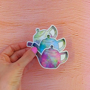 May include: Three colorful teapots with a galaxy print design. The teapots are stacked on top of each other. The teapots are a light green, blue, and purple.