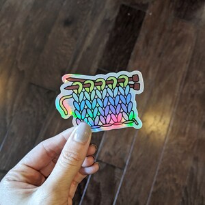 May include: A holographic sticker of a rainbow colored knitted fabric with two knitting needles. The fabric is in a stitch pattern that resembles a cable knit.