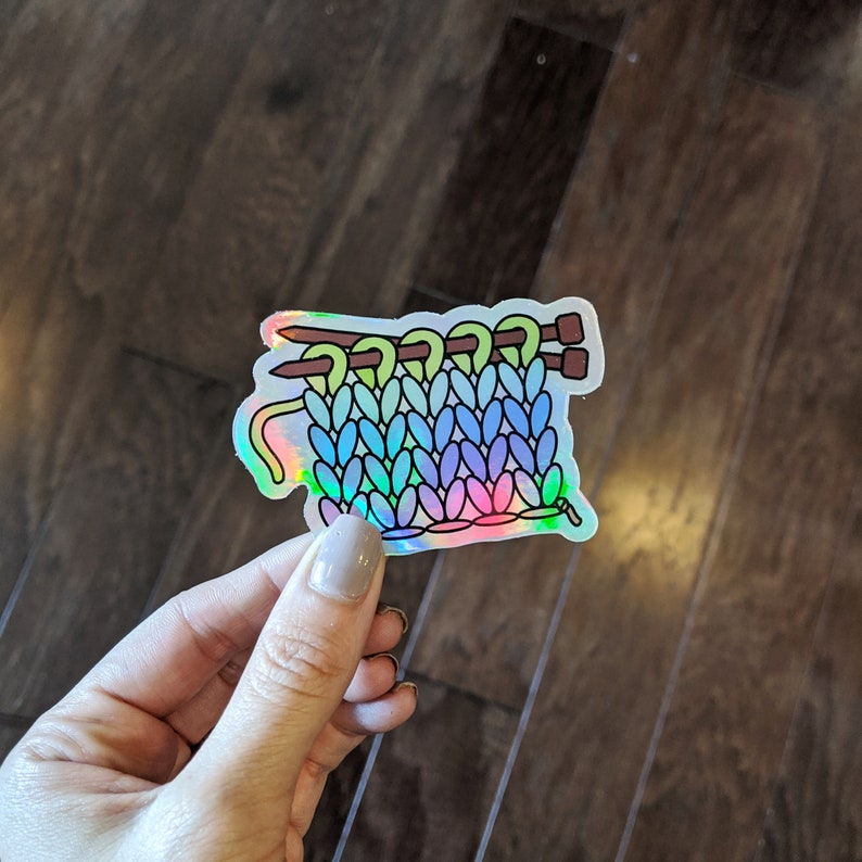 May include: A holographic sticker of a rainbow colored knitted fabric with two knitting needles. The fabric is in a stitch pattern that resembles a cable knit.