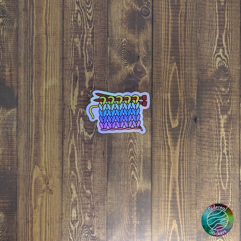 May include: A rainbow colored knit stitch sticker with two red knitting needles. The sticker is on a wood background.