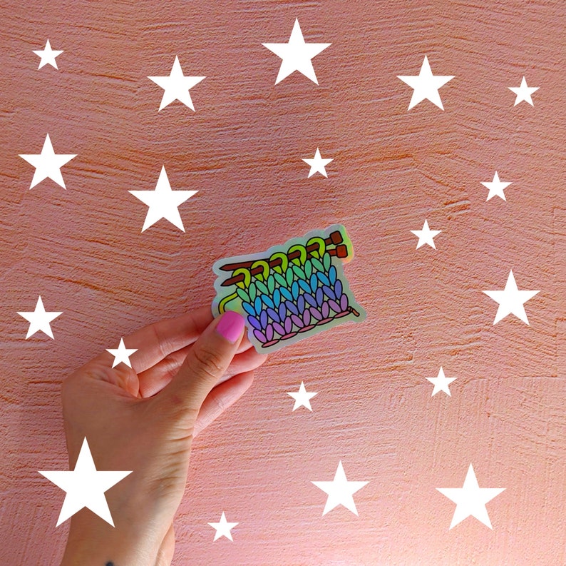 May include: A rainbow colored knitted fabric sticker with a brown knitting needle on a pink background with white stars.