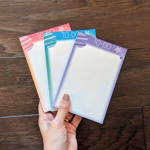 May include: Four colourful to-do list notepads with a space theme. Each notepad has a different colour and features a planet design with stars.