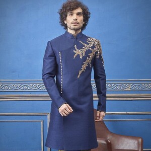 Puede incluir: Una kurta azul marino de manga larga con bordados dorados en el pecho. La kurta se combina con pantalones dorados y zapatos negros.