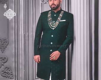 Puede incluir: Un sherwani verde oscuro para hombre con un collar de cuentas y pantalones blancos. El sherwani tiene una tela texturizada y está adornado con pequeñas incrustaciones plateadas. El hombre lleva mocasines negros.