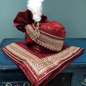 May include: A maroon turban with gold embroidery and a white feather. The turban is on a maroon fabric with gold embroidery and sequins.