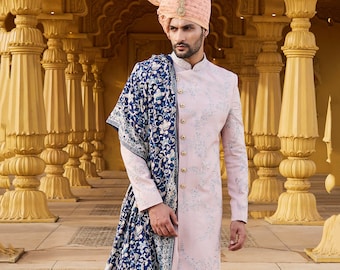 May include: A man wearing a pink and white patterned long-sleeved kurta with a blue and white patterned shawl draped over his shoulder. He is also wearing white pants and a pink turban with a white feather.