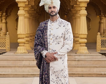 Könnte beinhalten: Ein weißer bestickter Sherwani mit einem blauen und goldenen Schal. Der Sherwani hat einen Stehkragen und einen Knopfverschluss. Der Schal ist über die linke Schulter gelegt und mit komplizierten Blumenmustern verziert.