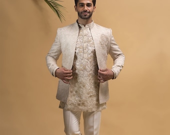May include: A man wearing a white embroidered jacket, a white embroidered shirt, white pants, and brown shoes. The man is standing with one leg crossed in front of the other.