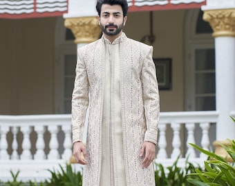 Puede incluir: Un hombre lleva un sherwani bordado de color crema con un frente largo y abierto y pantalones a juego. Está de pie frente a un edificio con una barandilla blanca y plantas verdes.