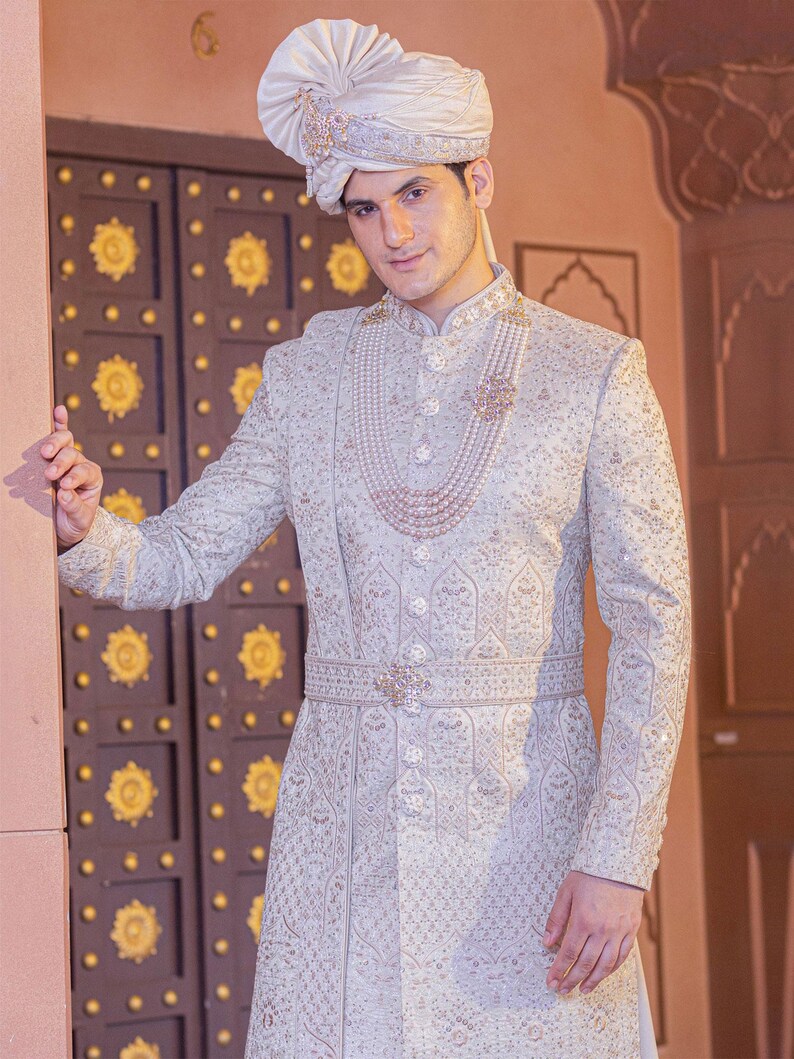 May include: A light blue embroidered sherwani with a matching turban. The sherwani features intricate patterns, a pearl necklace, and a decorative belt. The outfit is worn with a matching turban, creating a formal and elegant look.