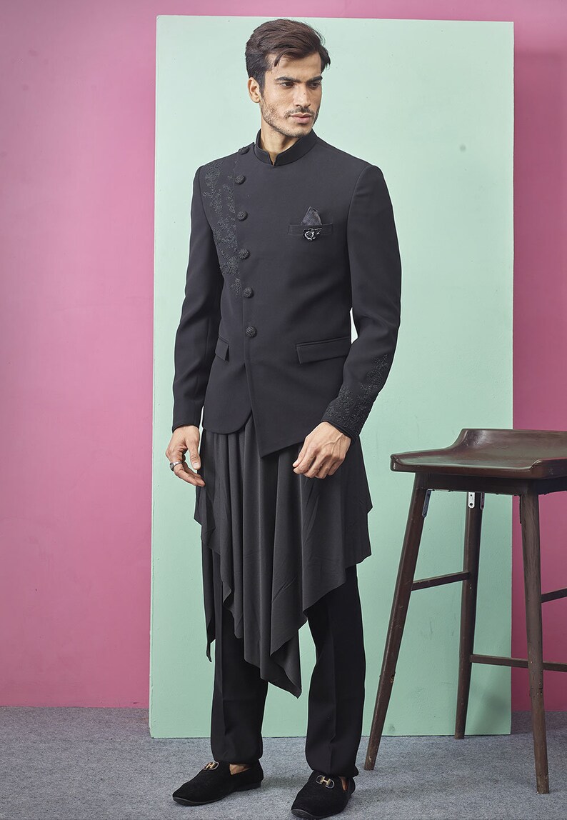 May include: Black formal suit with a mandarin collar jacket featuring embroidered detailing on the lapel and sleeves. The suit includes a draped asymmetrical top and black trousers. The model is wearing black loafers.