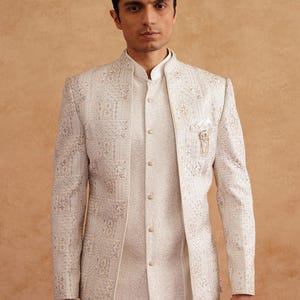 May include: A cream-colored, intricately patterned jacket and matching vest with a mandarin collar. The jacket features a detailed, embossed design and a small pocket square. The outfit is paired with white pants, creating a formal ensemble.