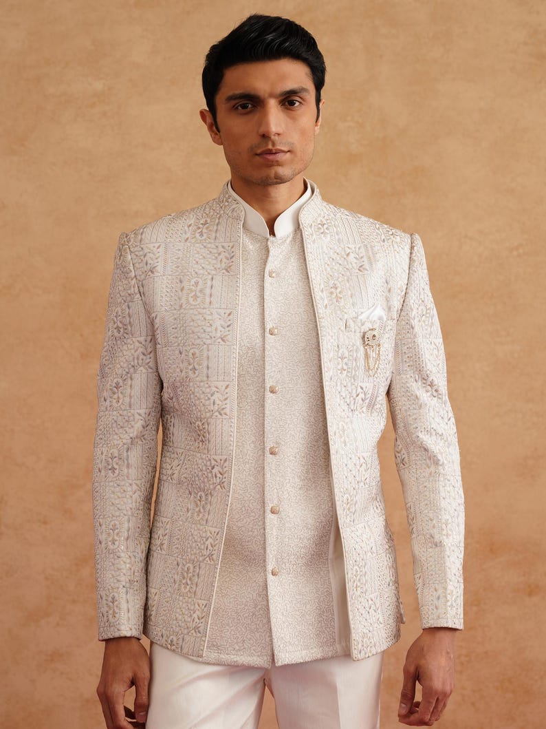 May include: A cream-colored, intricately patterned jacket and matching vest with a mandarin collar. The jacket features a detailed, embossed design and a small pocket square. The outfit is paired with white pants, creating a formal ensemble.