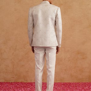 May include: A cream-colored, patterned blazer and white trousers. The blazer features an intricate design and a mandarin collar. The trousers are straight-legged. The person is standing on a pink surface.