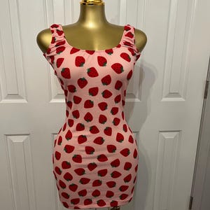 May include: A pink, ribbed mini dress with a repeating pattern of red strawberries. The dress has a scoop neckline and ruffled straps. The dress is displayed on a gold mannequin.