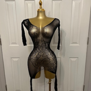 May include: Black fishnet lingerie with a floral pattern, displayed on a gold mannequin. The garment features long sleeves and a low neckline. The lingerie is designed with a cutout on the lower portion and attached straps.