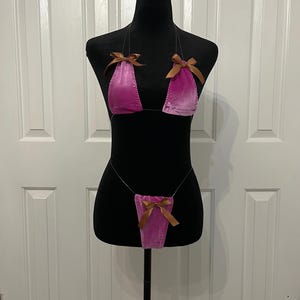 May include: A pink velvet bikini set with brown satin bows. The top has triangle cups and the bottom is a thong style. The set is displayed on a black mannequin against a white door background.