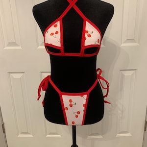May include: A red and white cherry print bikini with a halter top and a high-cut bottom. The bikini has red trim and ties at the neck and sides.