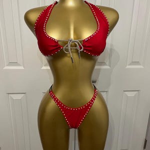 May include: Red bikini set with a halter top and thong-style bottoms. The bikini is adorned with small, sparkling embellishments along the edges and a silver-toned tie detail at the bust. The set is displayed on a gold mannequin.