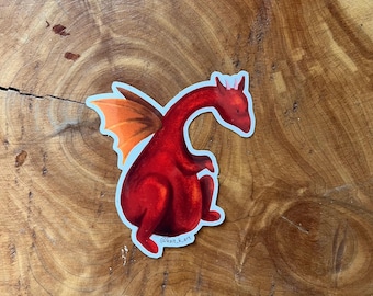 Könnte beinhalten: Ein roter Drachen-Aufkleber mit orangefarbenen Flügeln. Der Drache sitzt auf seinen Hinterbeinen und hat den Kopf zur Seite gedreht. Der Aufkleber hat einen weißen Rand und den Text "@kait.k.art" in der rechten unteren Ecke.